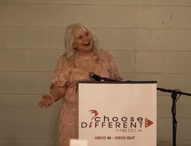 Woman with blonde hair in pink dress speaking at a podium with a microphone, smiling, against a plain wall.