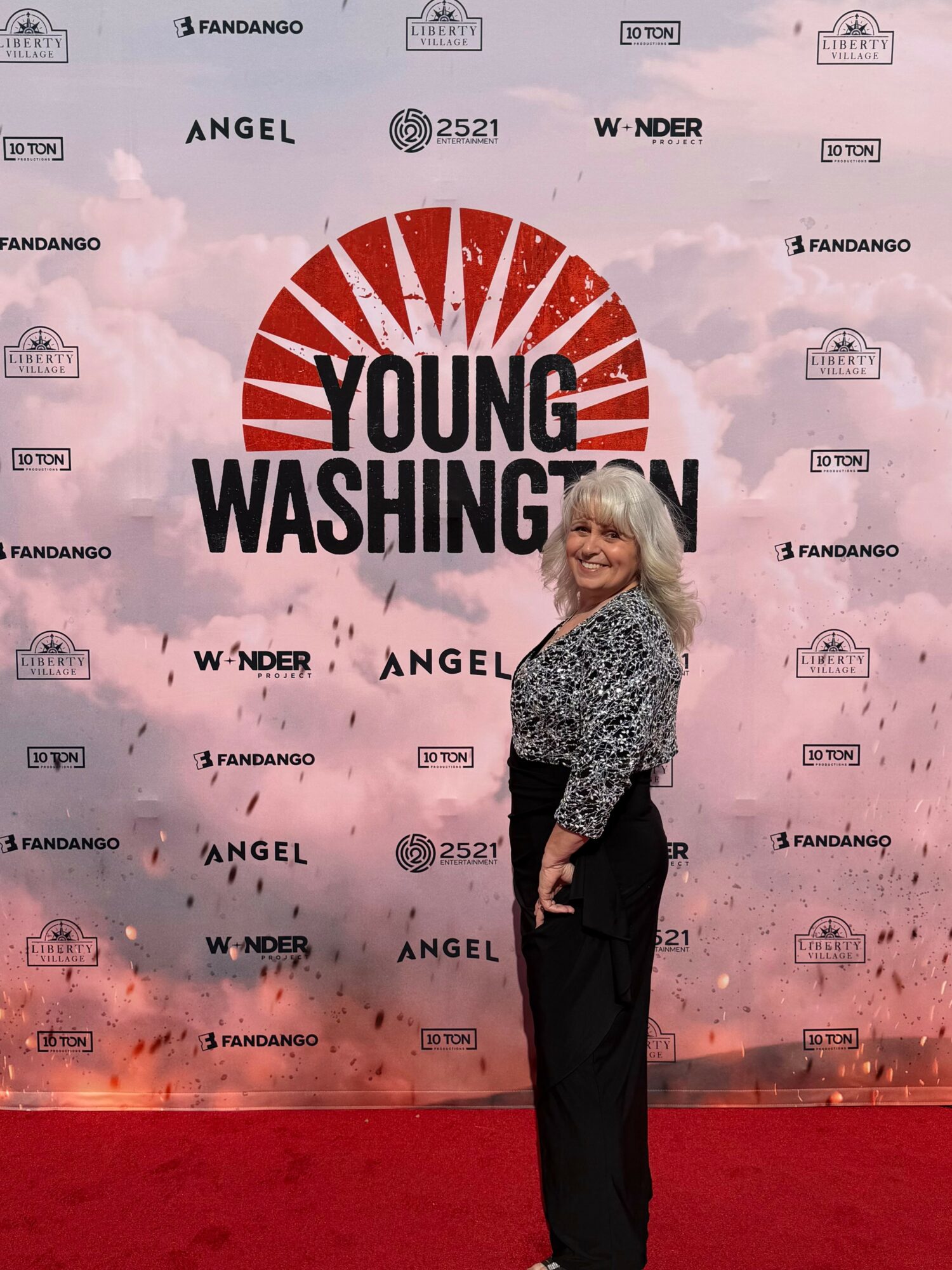 Woman with blonde hair standing on red carpet in front of a pink backdrop with a red sunburst and text, smiling.