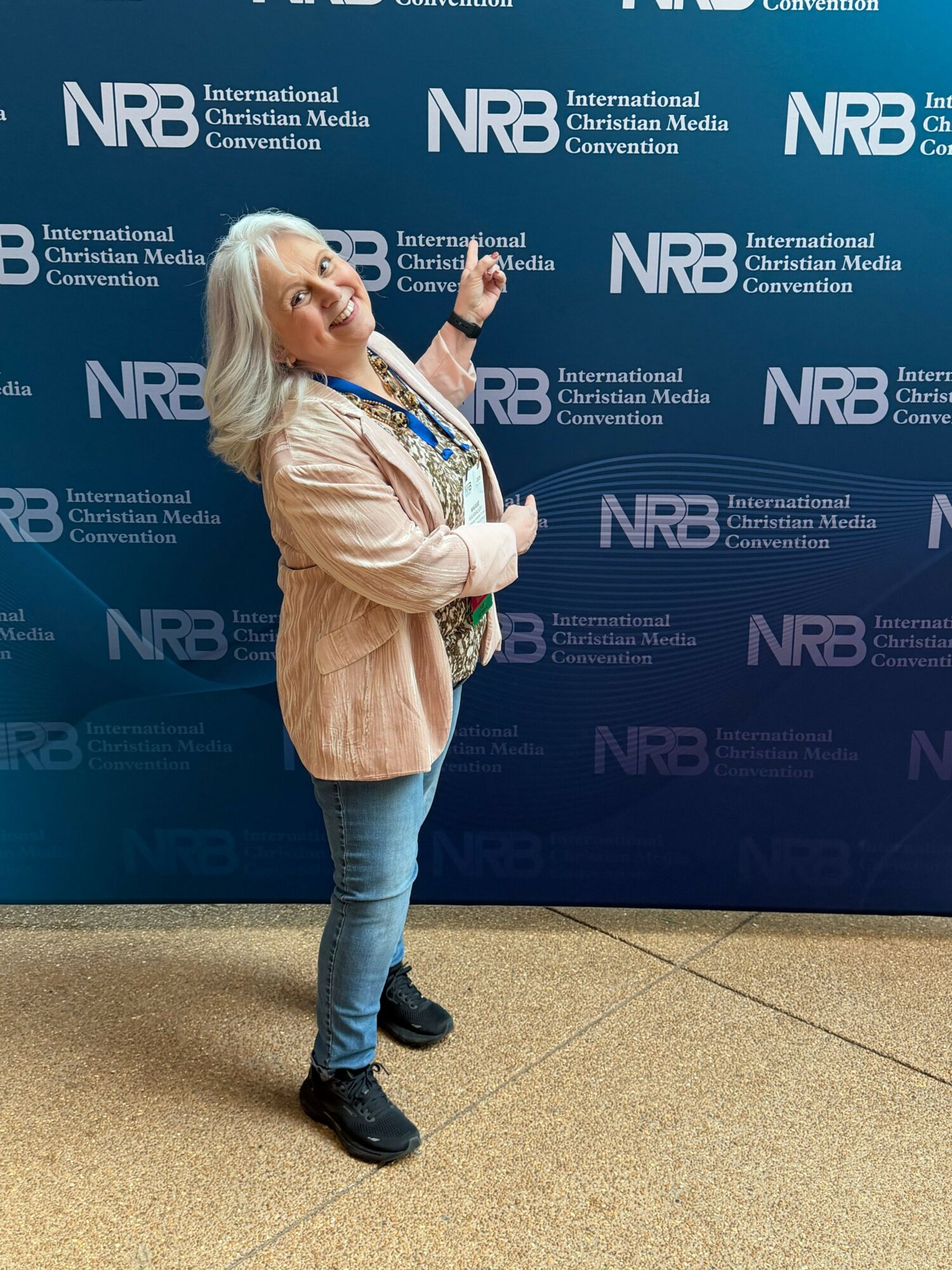 Woman with blonde hair standing sideways, pointing at a blue NRB backdrop, wearing a beige coat, jeans, and black boots.
