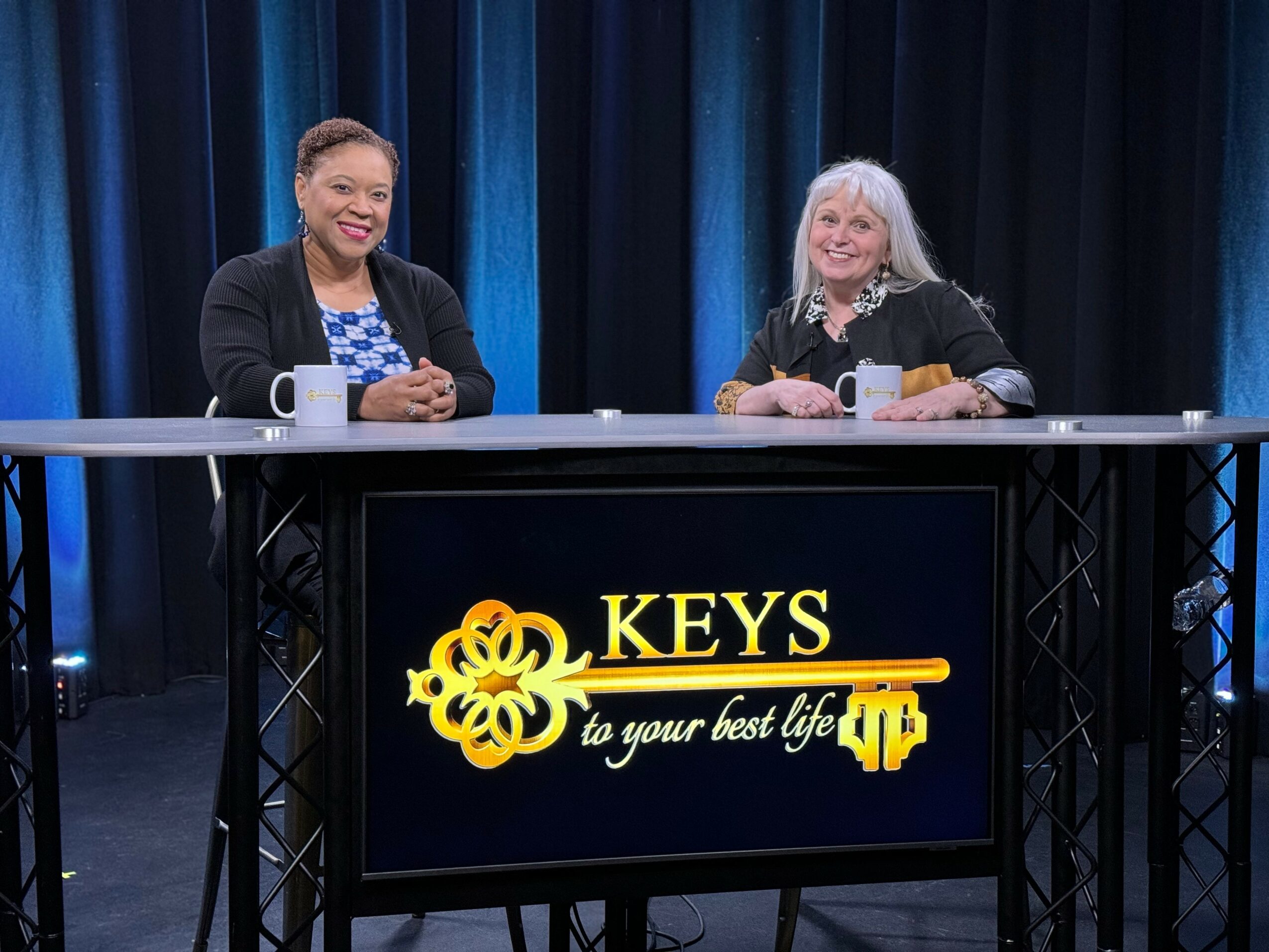 Two women sit at a table with a screen displaying the logo and text, in front of a dark blue curtain backdrop.