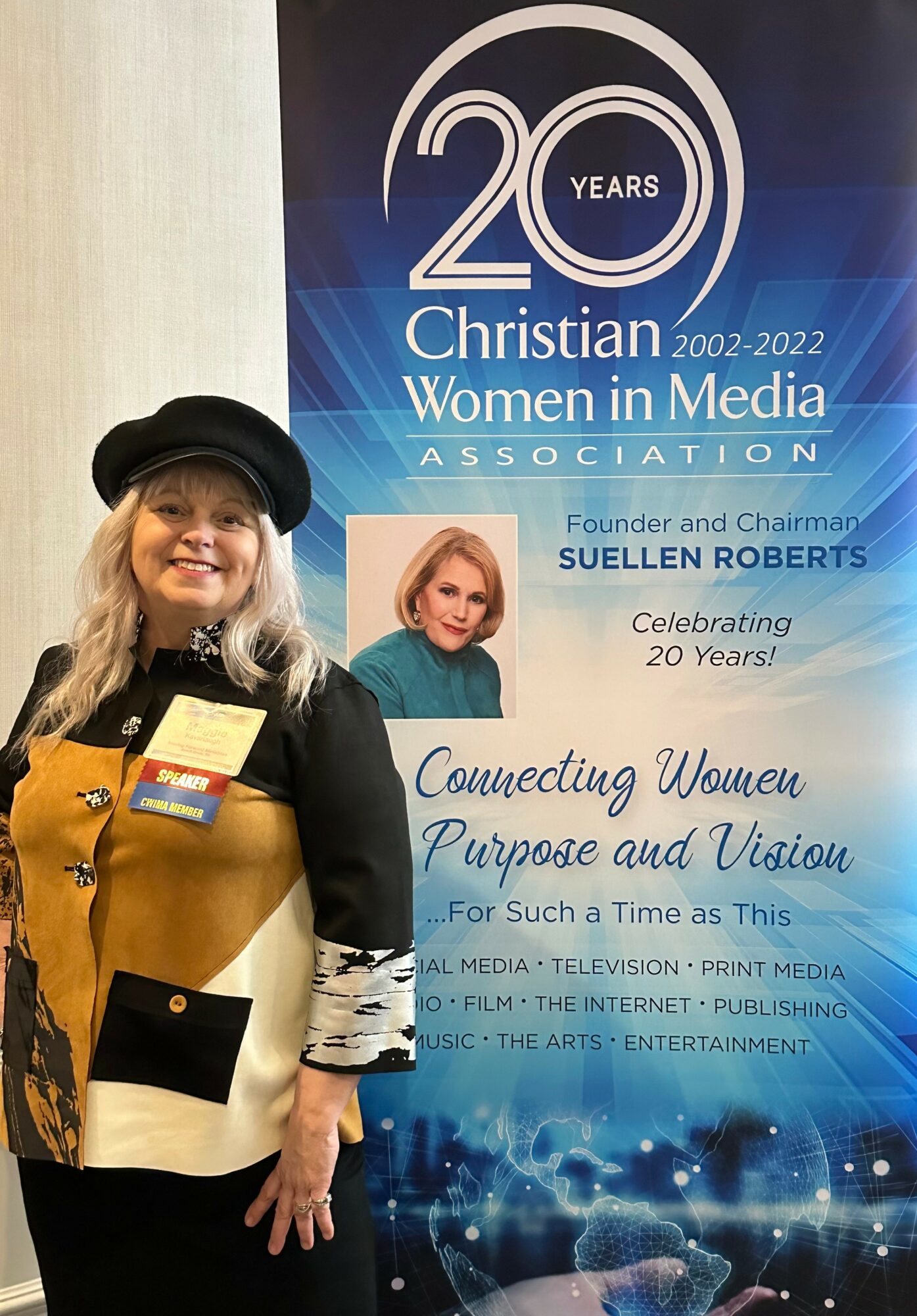 Woman with blonde hair wearing a black hat and coat, holding a yellow bag, standing in front of a blue banner celebrating 20 years of Women in Media.