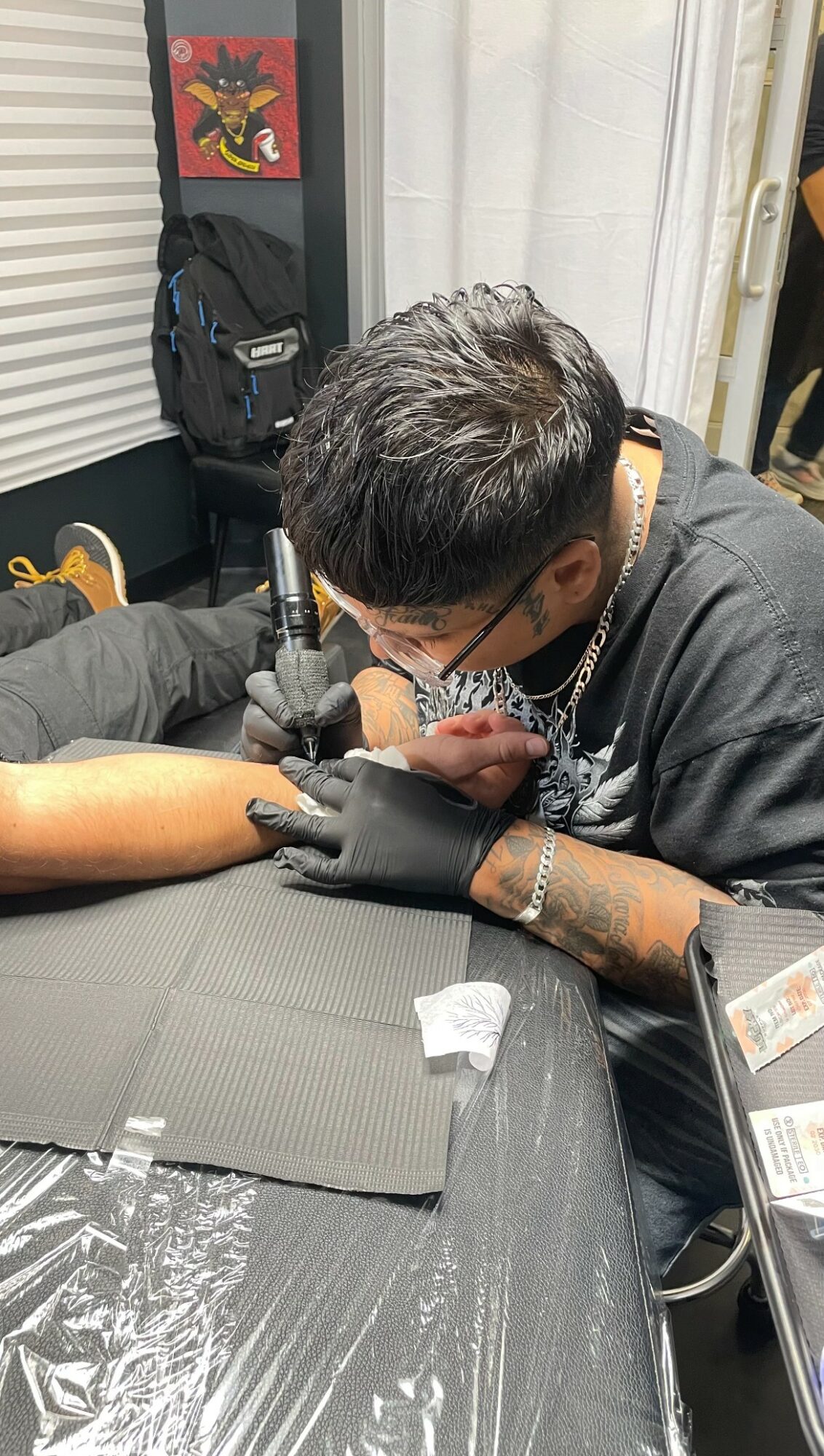 Person receiving a tattoo on their arm in a tattoo studio, with equipment and supplies visible around.