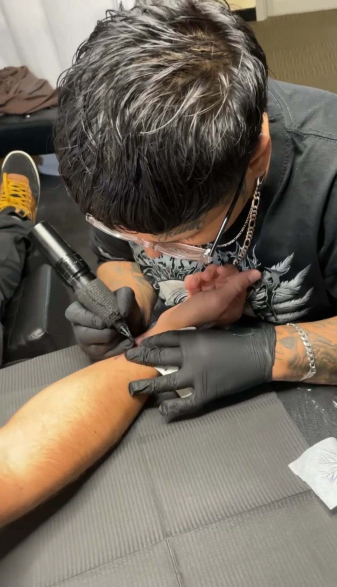 Tattoo artist working on a person's arm with tattoo equipment, wearing black gloves, in a studio setting.