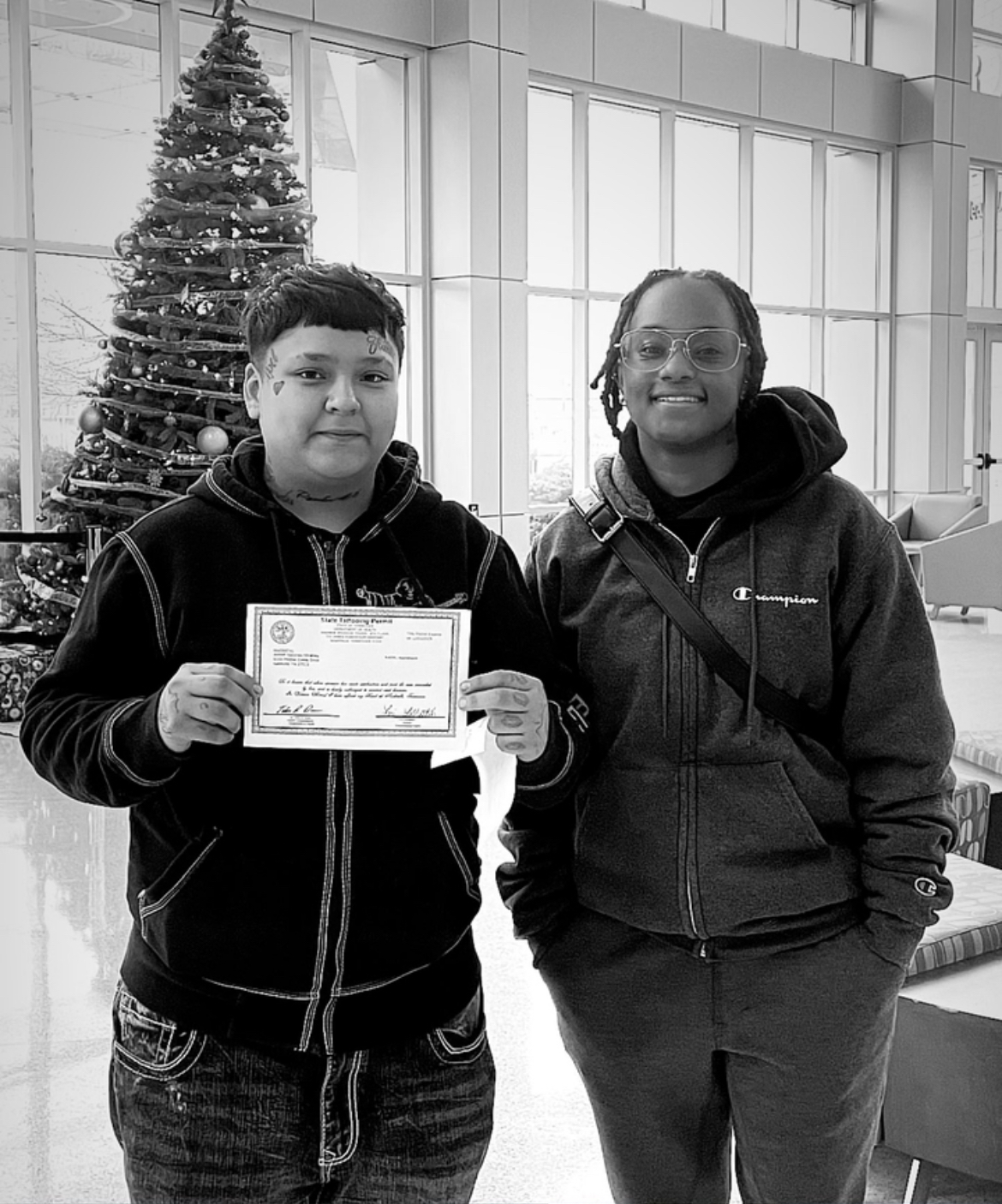 Two young people standing indoors near a decorated Christmas tree, one holding a certificate, smiling at the camera.