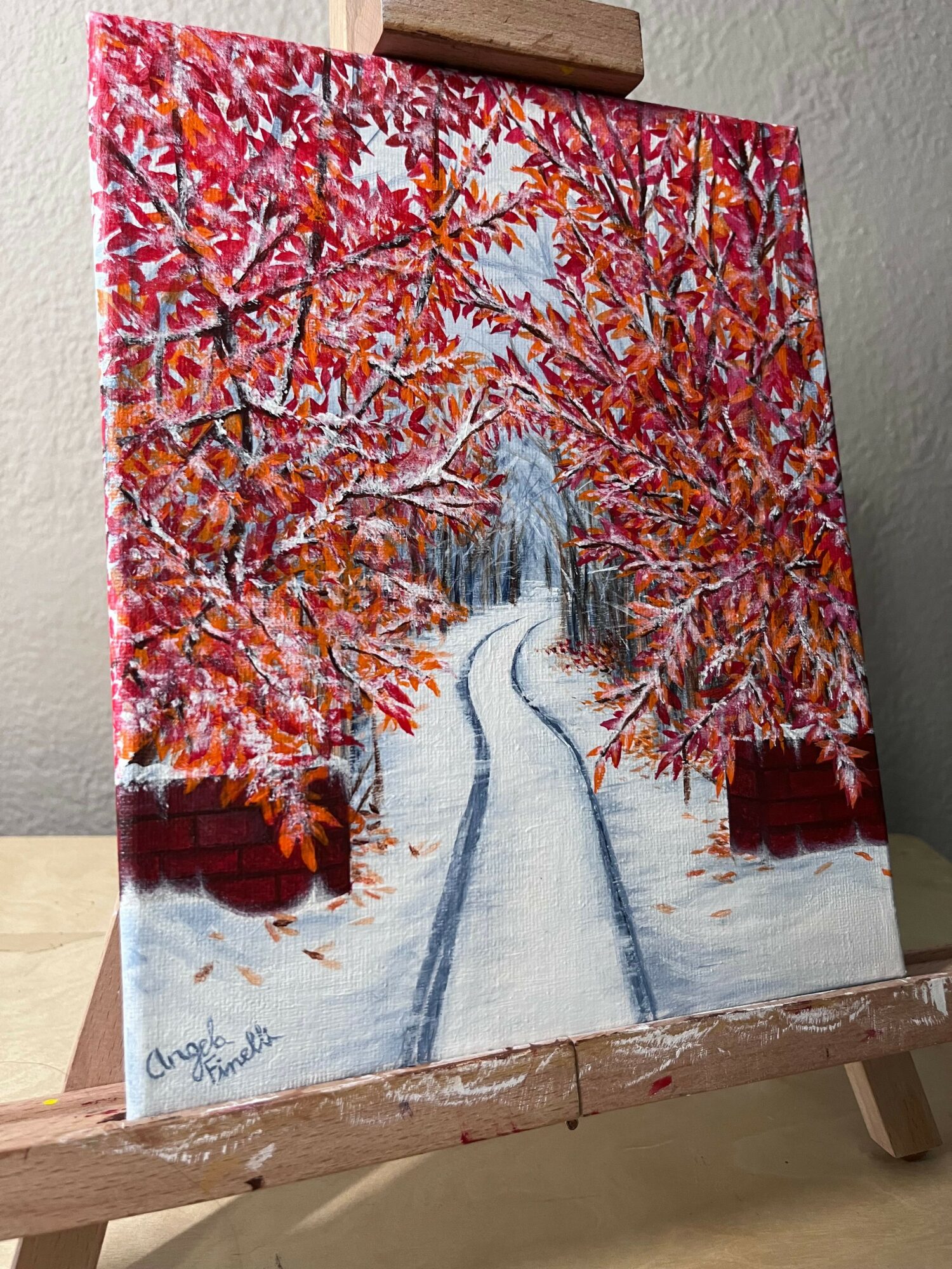 Painting of a tree with red and orange leaves beside a body of water, with a reflection and a signature in the corner.
