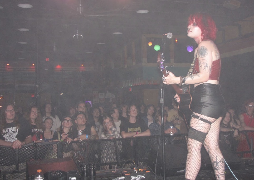 Performer with red hair, tattoos, and black outfit on stage, audience watching in background, dim lighting, colorful stage lights.