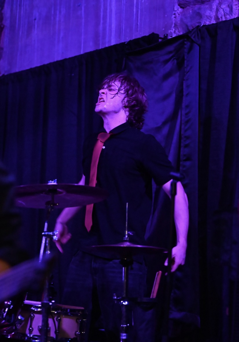 Person with curly hair stands on stage with a black curtain background, smiling with eyes closed, wearing a black jacket and red shirt.