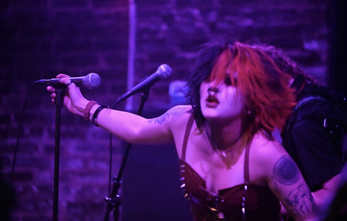Performer with red and black hair singing into a microphone on stage with purple lighting.