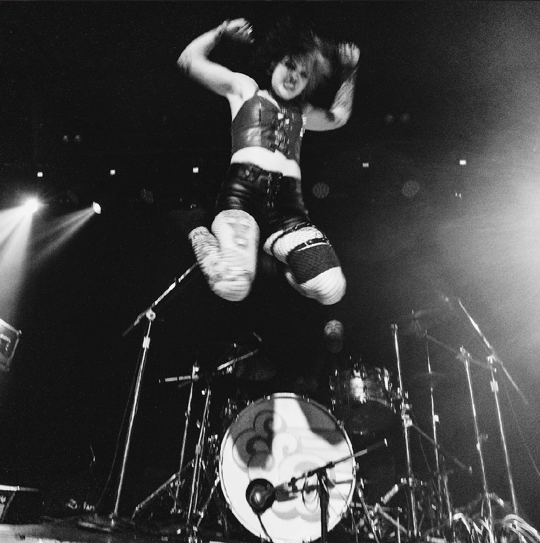 Performer jumps in front of a drum set on stage, with stage lights shining around. Black and white image.