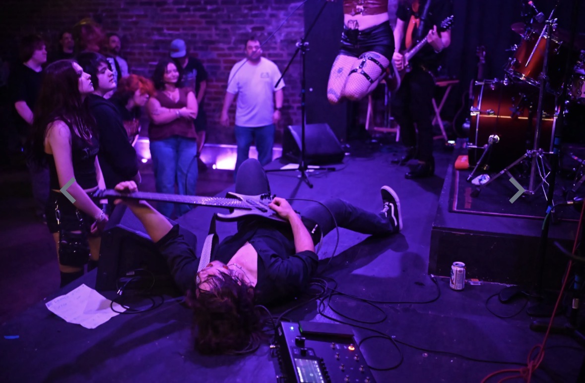 Person lying on stage with a guitar, surrounded by an audience and band members, in a music venue.