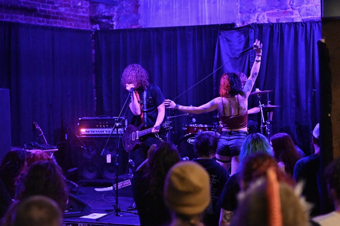 Musicians perform on stage with a guitarist and a singer, audience watching in a dimly lit venue.