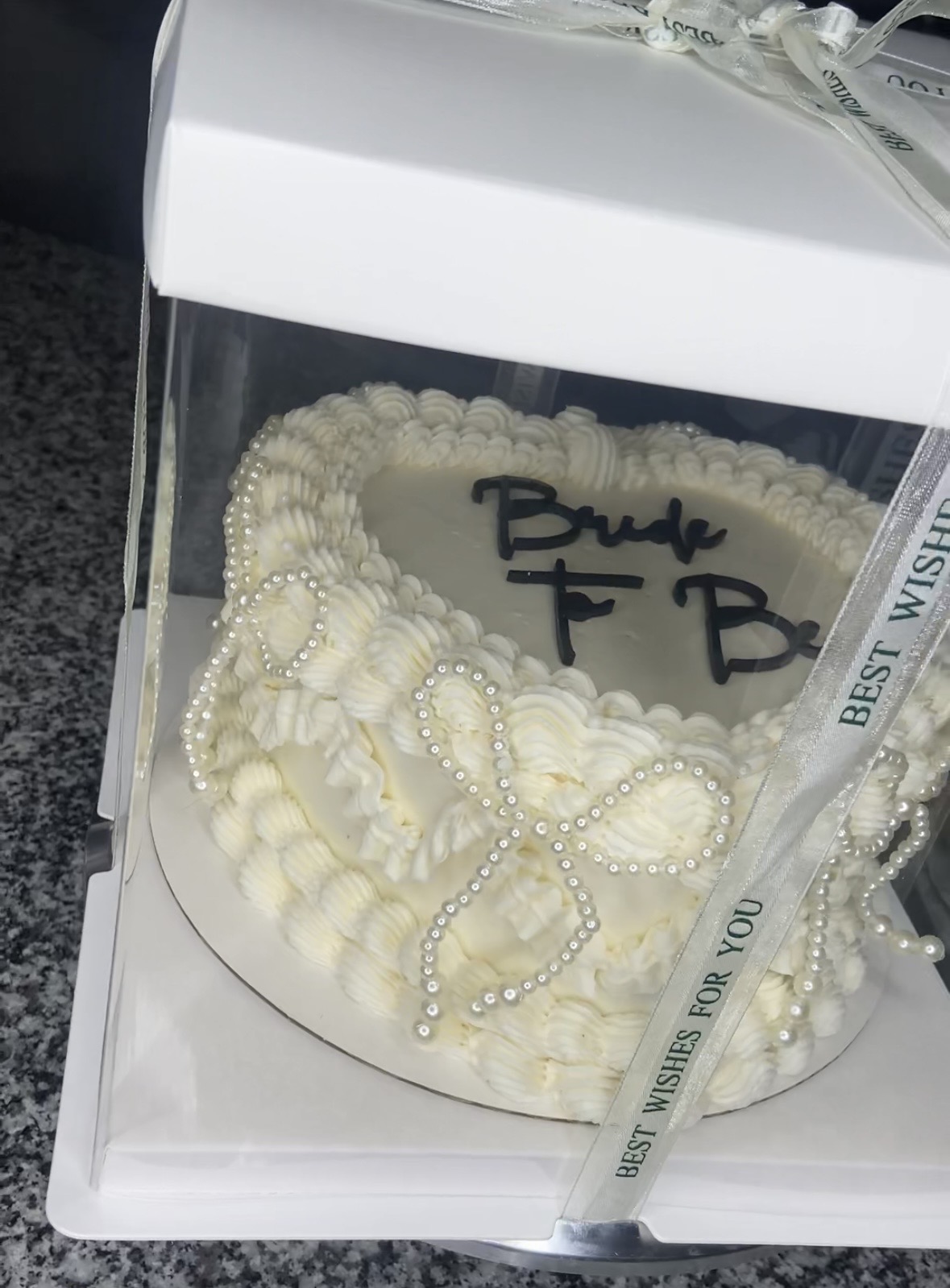 Heart-shaped cake decorated with white pearls and cream, inside a clear display box, with handwritten message on top.