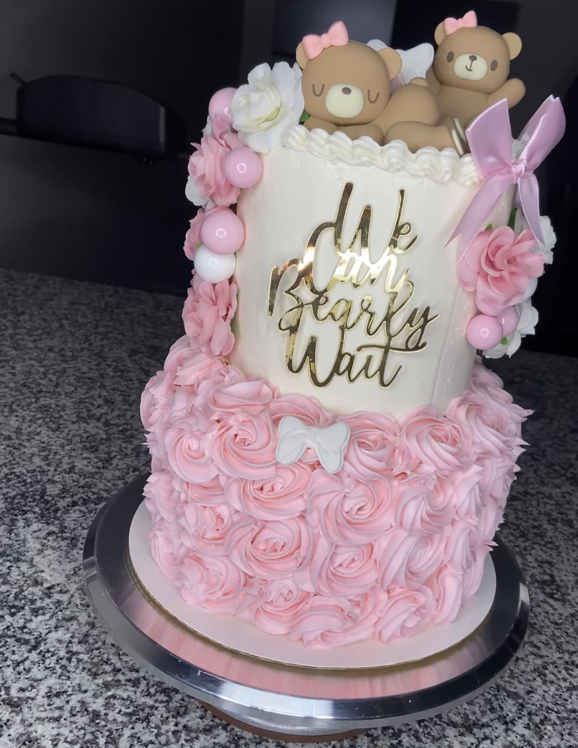 Decorative cake with pink rosette frosting, two bear figurines on top, pink bows, and a message in gold script.