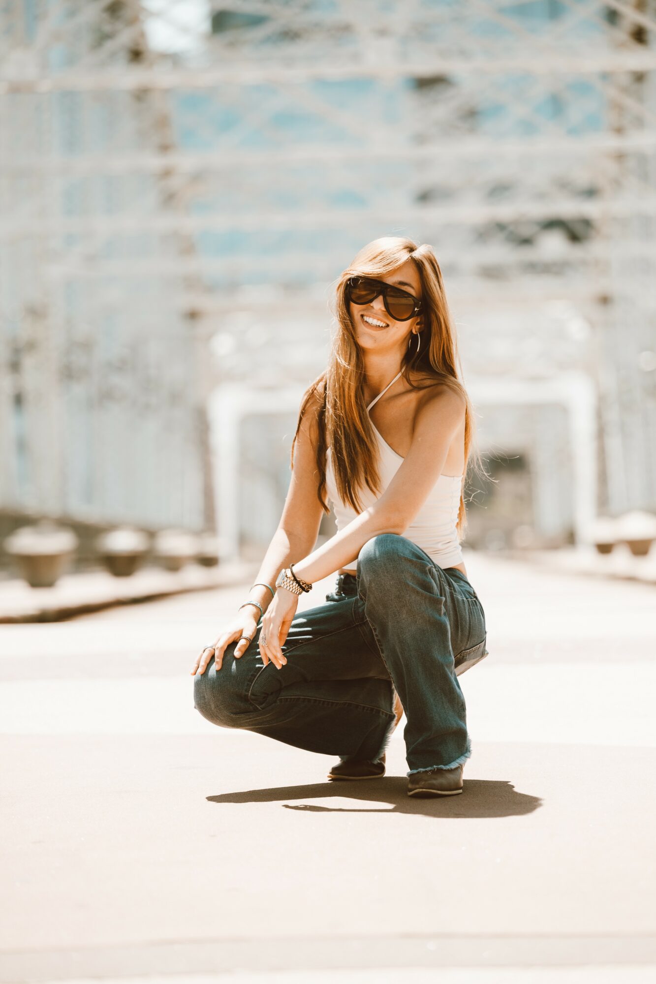 Young woman with long hair, sunglasses, white top, and jeans crouching outdoors on a sunny day.