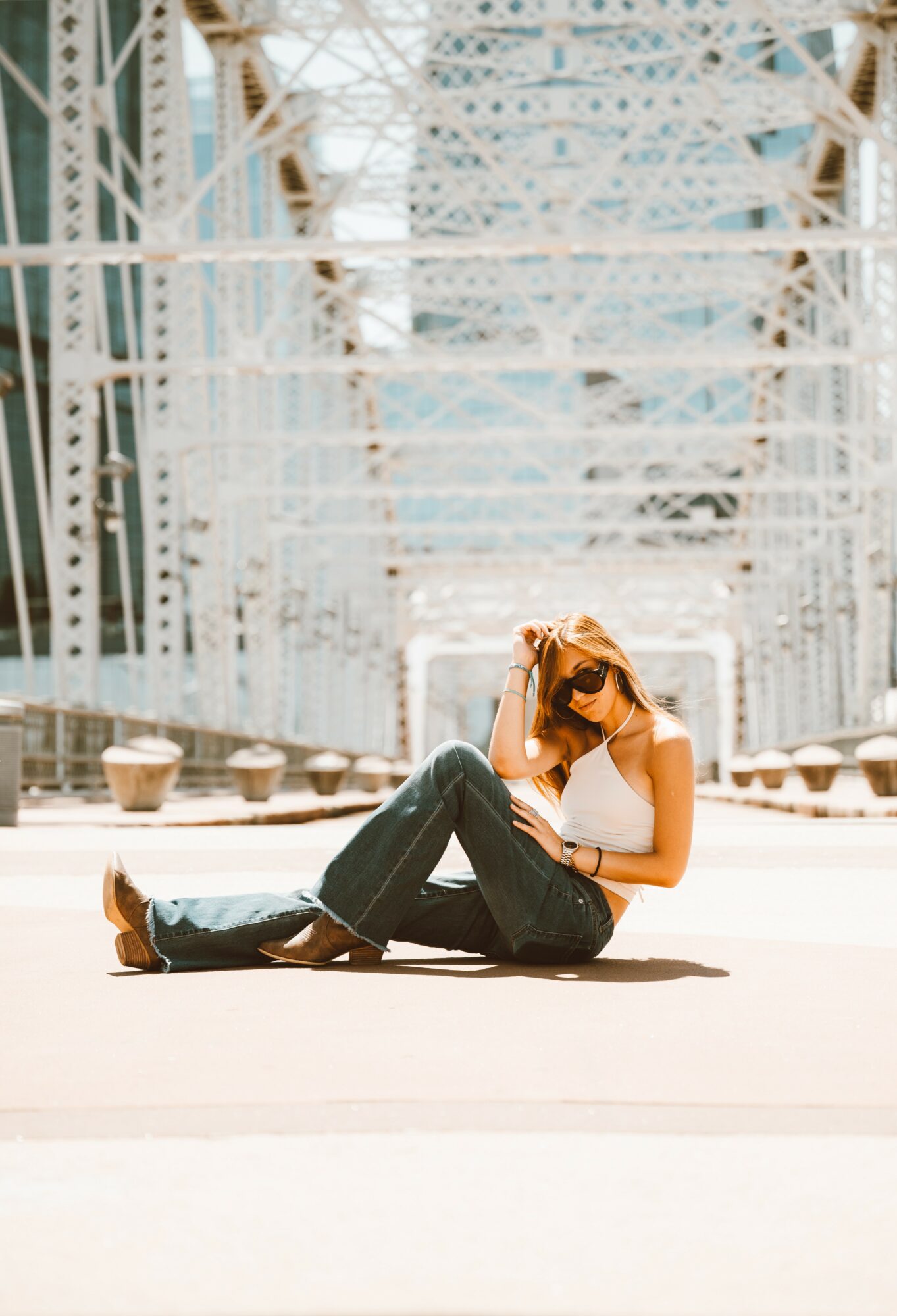 Woman sitting on ground, holding sunglasses, in front of a large bridge structure with metal beams and cables.