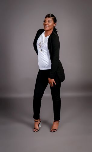 Woman standing with hands on hips, smiling, wearing a white top, black jacket, black pants, and high heels.