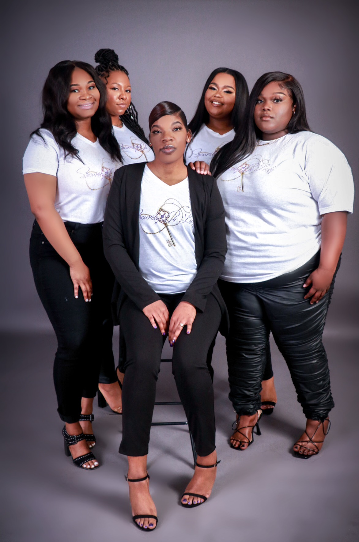 Group of five women standing together, posing for a photo against a gray background, dressed in casual and semi-formal clothing.