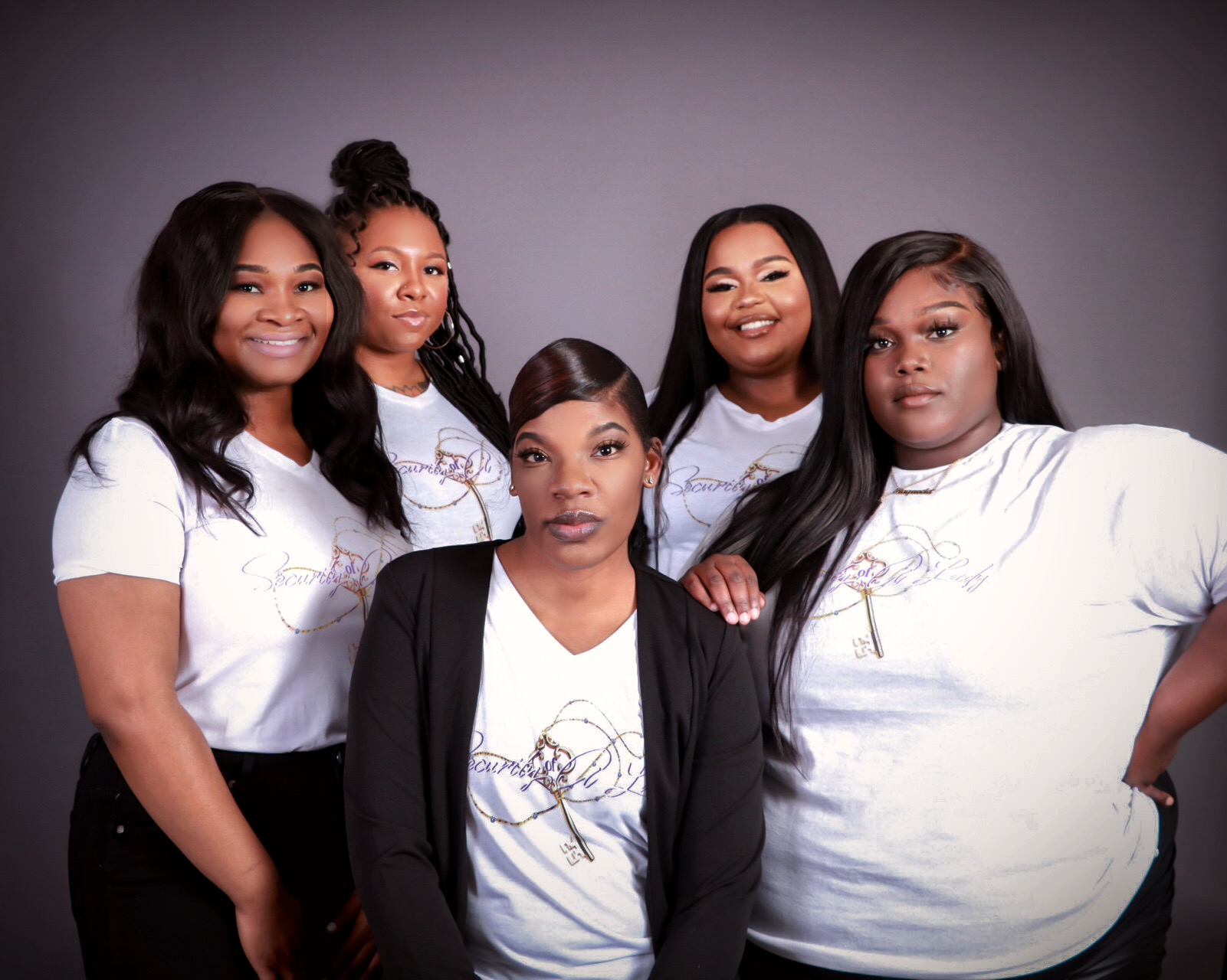 Group of five women standing together, smiling, against a plain background.