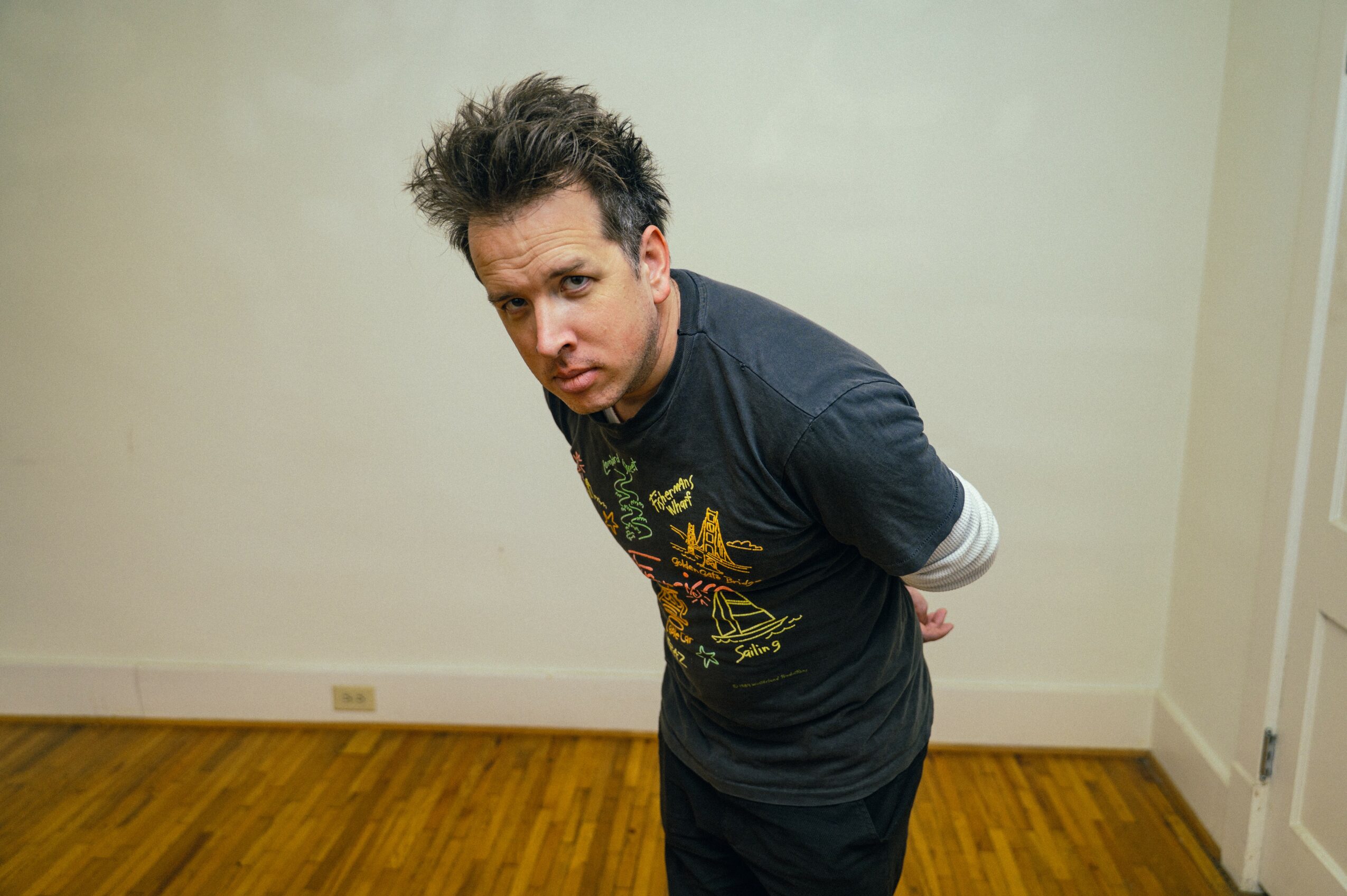 Person with spiky hair leaning forward, hands behind back, in a room with wooden floor and white walls.