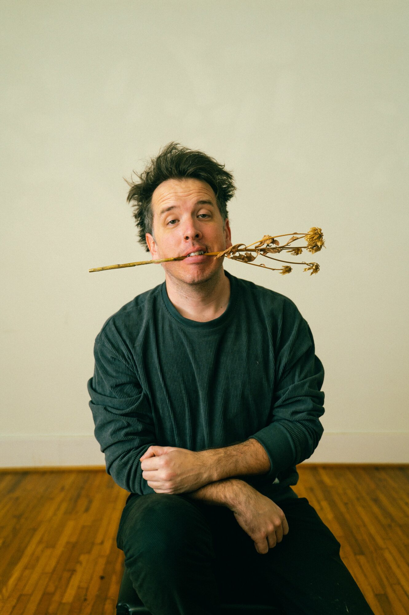 Person with messy hair holding a stick with a knot in their mouth, sitting on a wooden floor against a plain wall.