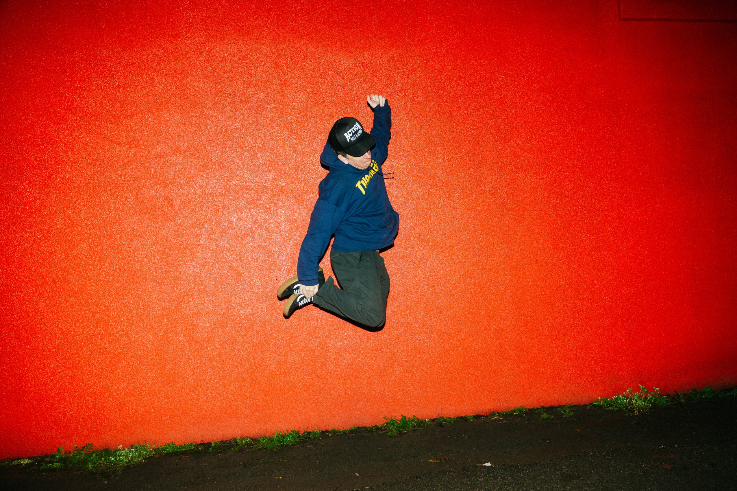 Person in blue hoodie and gray pants jumping against a red wall, with grass and pavement below, raising one arm.