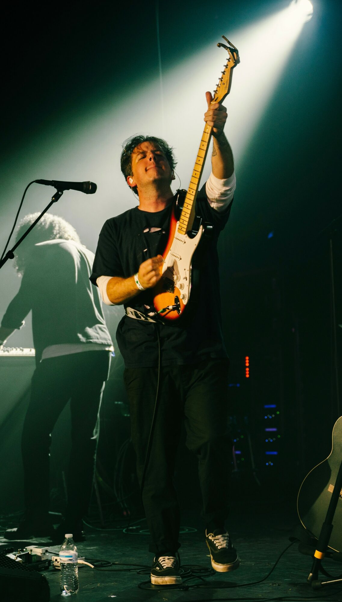 Person playing electric guitar on stage with a microphone and shadow in background.
