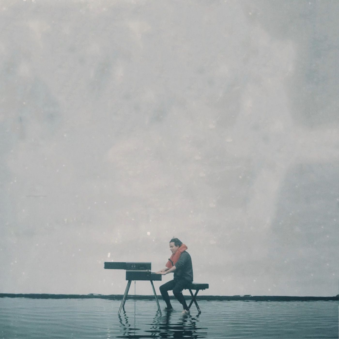 Person sitting on a stool playing a keyboard outdoors against a cloudy sky, with water in the background.
