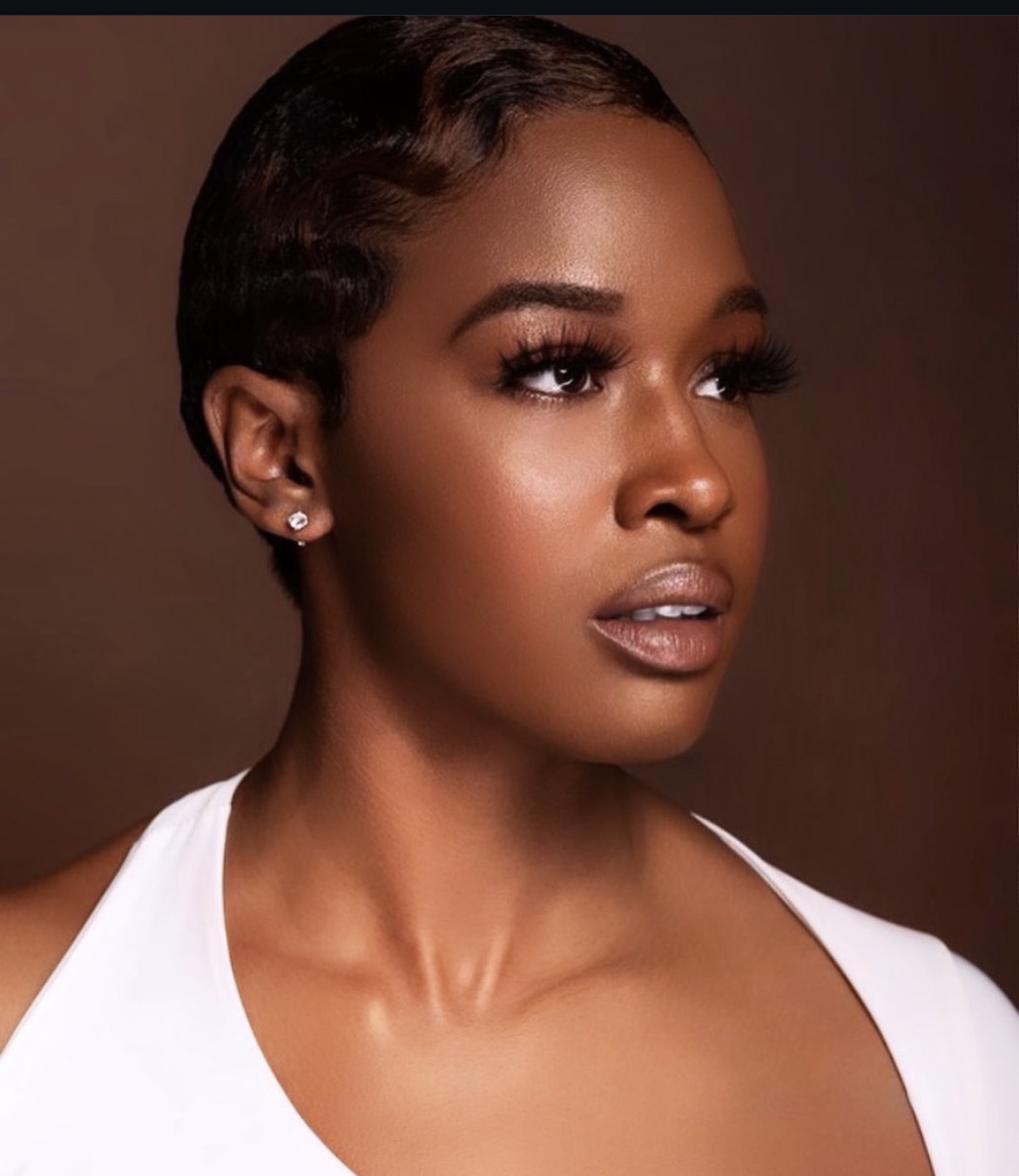 Portrait of a woman with short, wavy hair, wearing earrings and a white top, looking to the side against a dark background.
