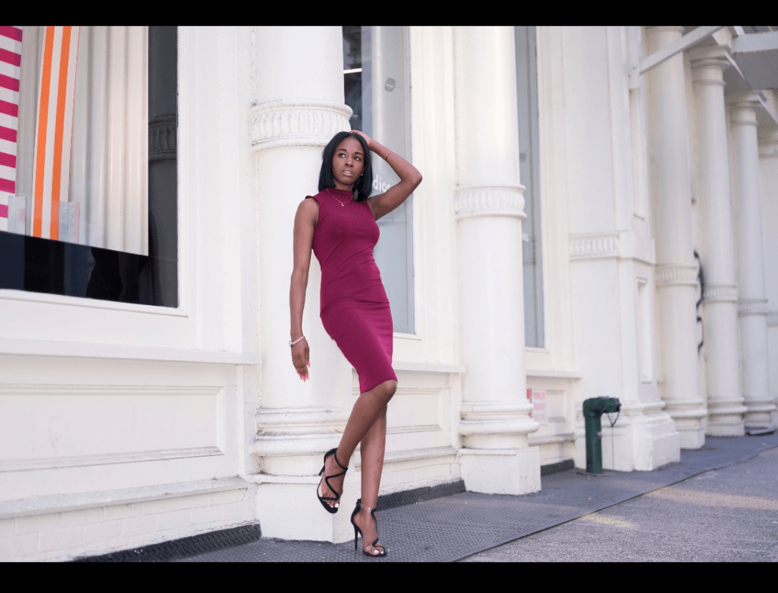 Woman in a pink dress and high heels standing against white columns, posing with one hand on her head.