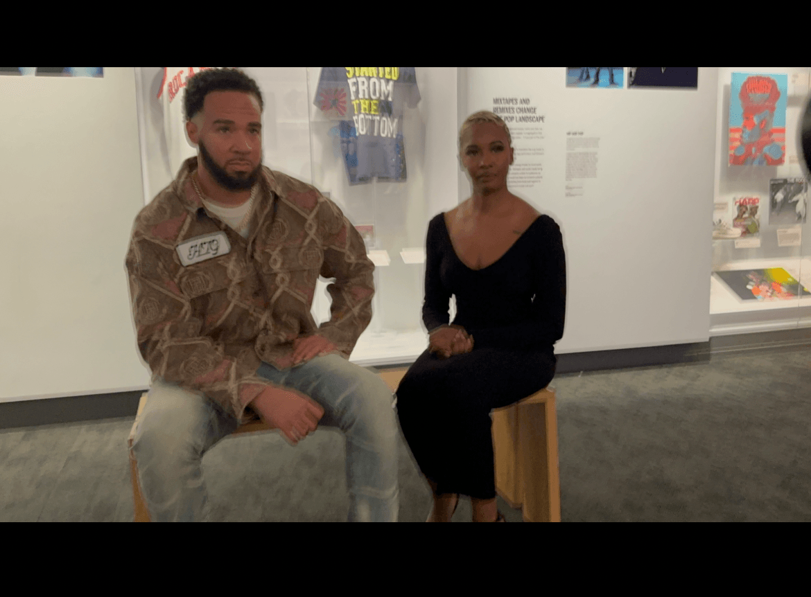 Two people sitting on stools in an art gallery, with posters and artwork on the walls behind them.