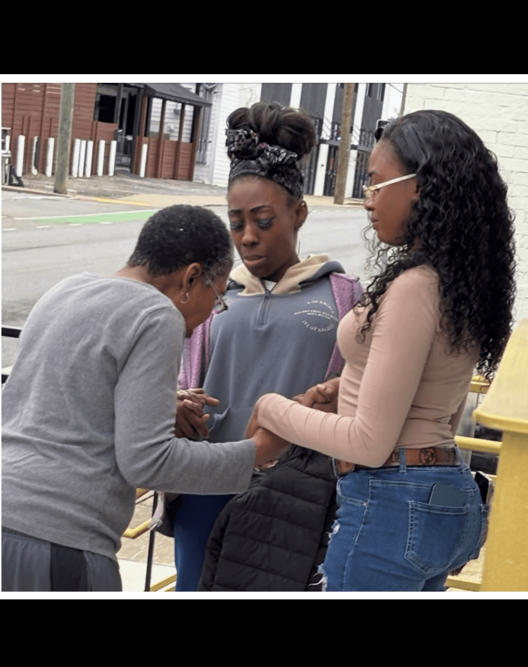 Three women standing outdoors, engaged in conversation, with buildings and street in background.