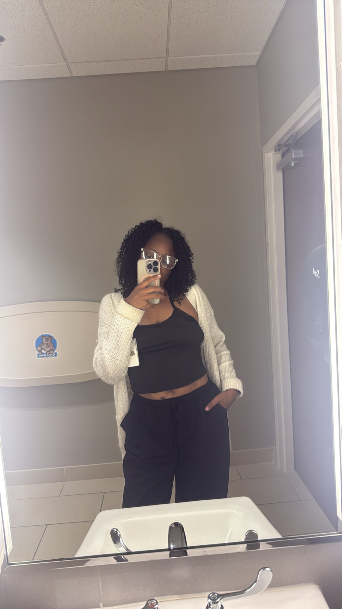 Person taking a mirror selfie in a room with a water fountain, wearing a black top, beige cardigan, and dark pants.