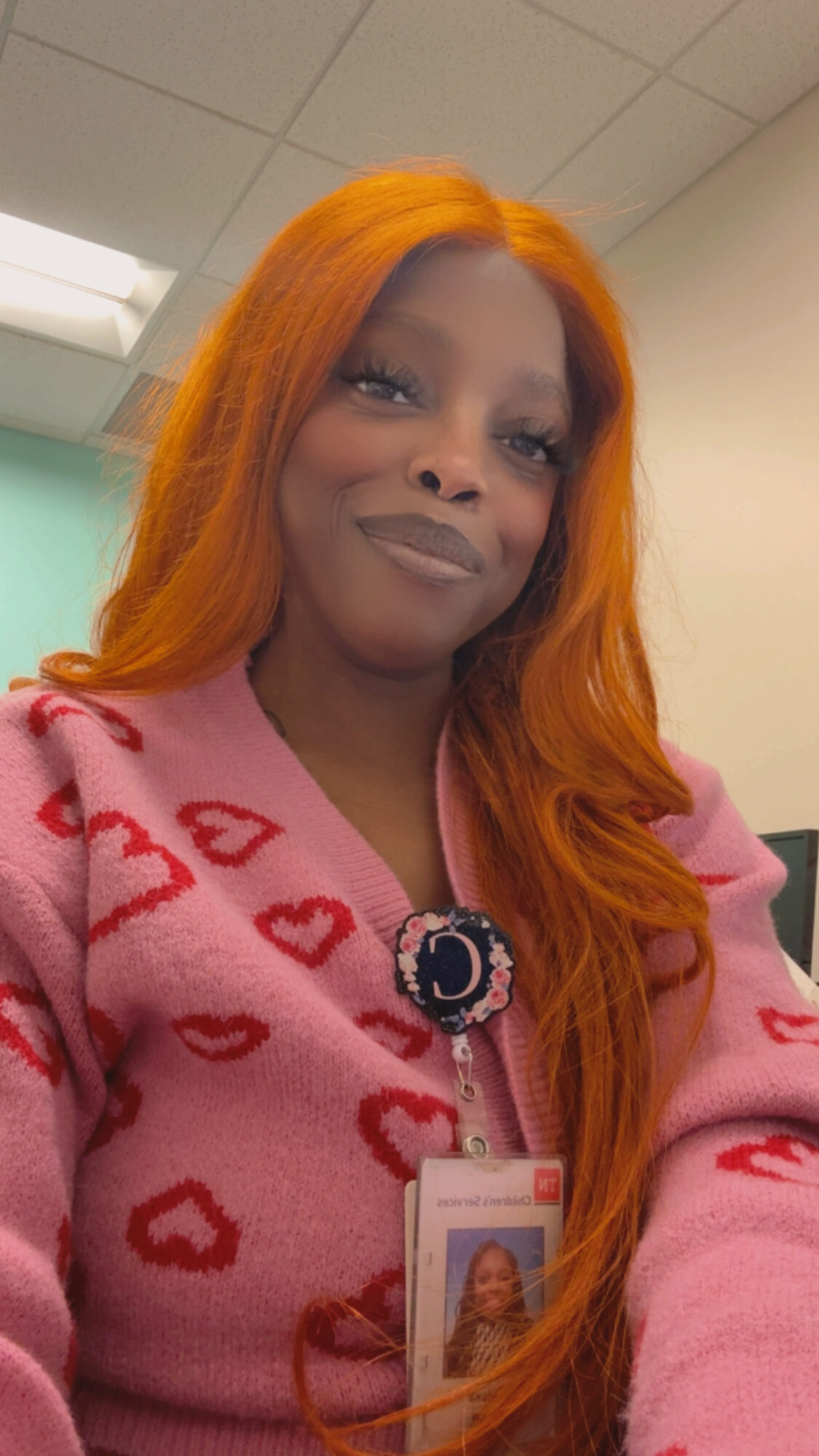 Woman with long orange hair wearing pink sweater with red hearts, smiling, in an indoor setting.