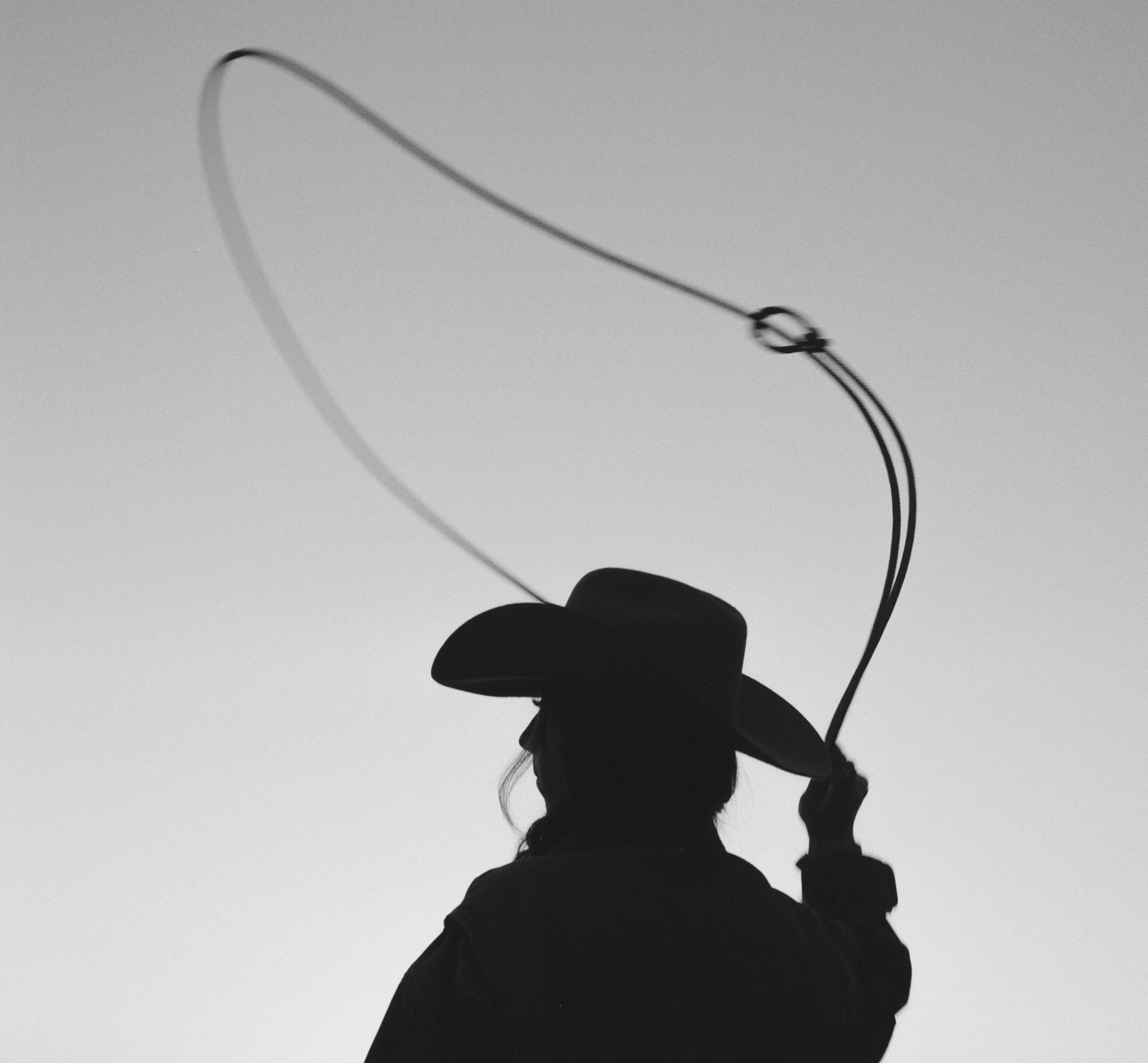 Silhouette of a person wearing a hat, holding a lasso in the air against a plain background.