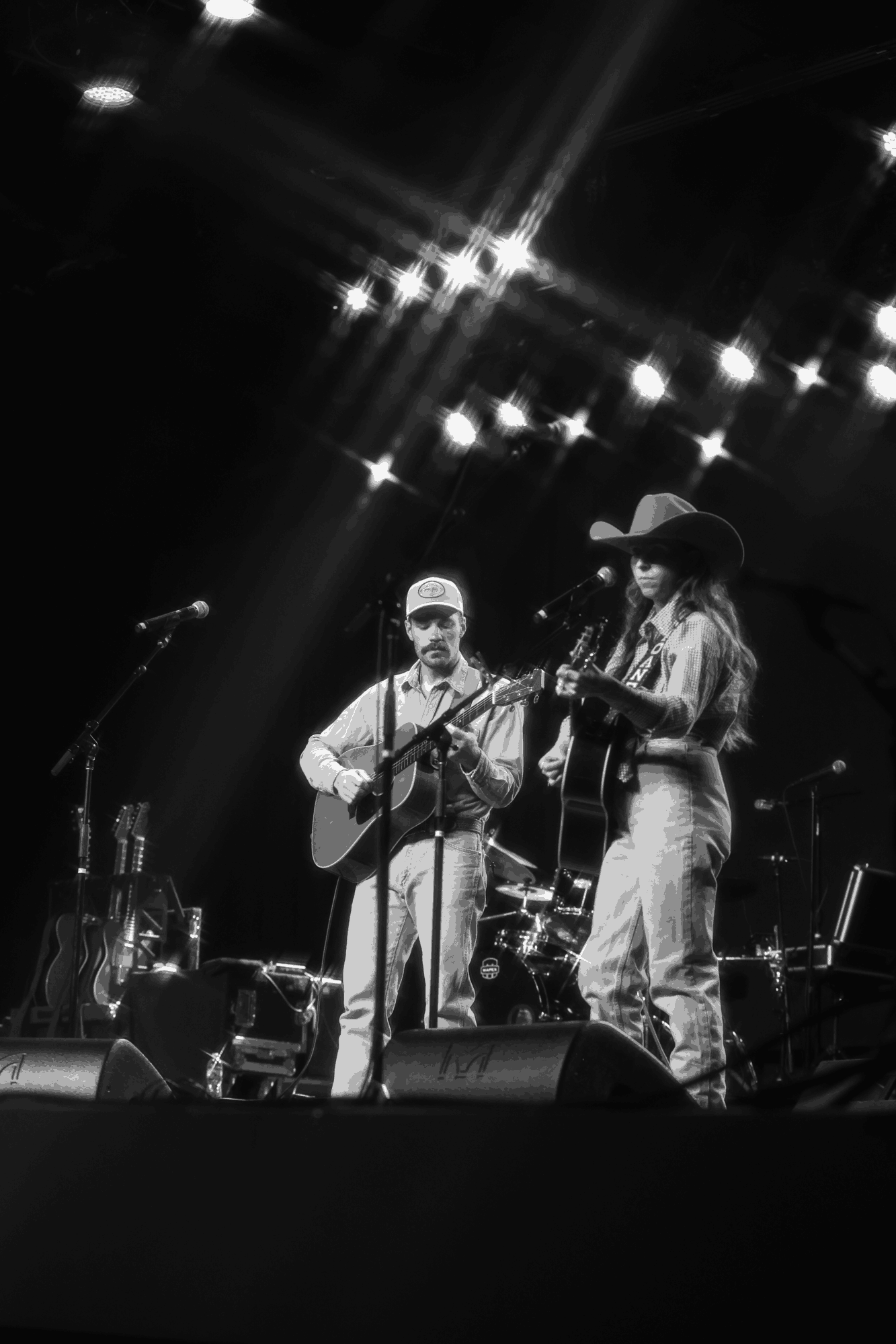 Two musicians perform on stage with guitars, one wearing a hat, under bright stage lights, in black and white.