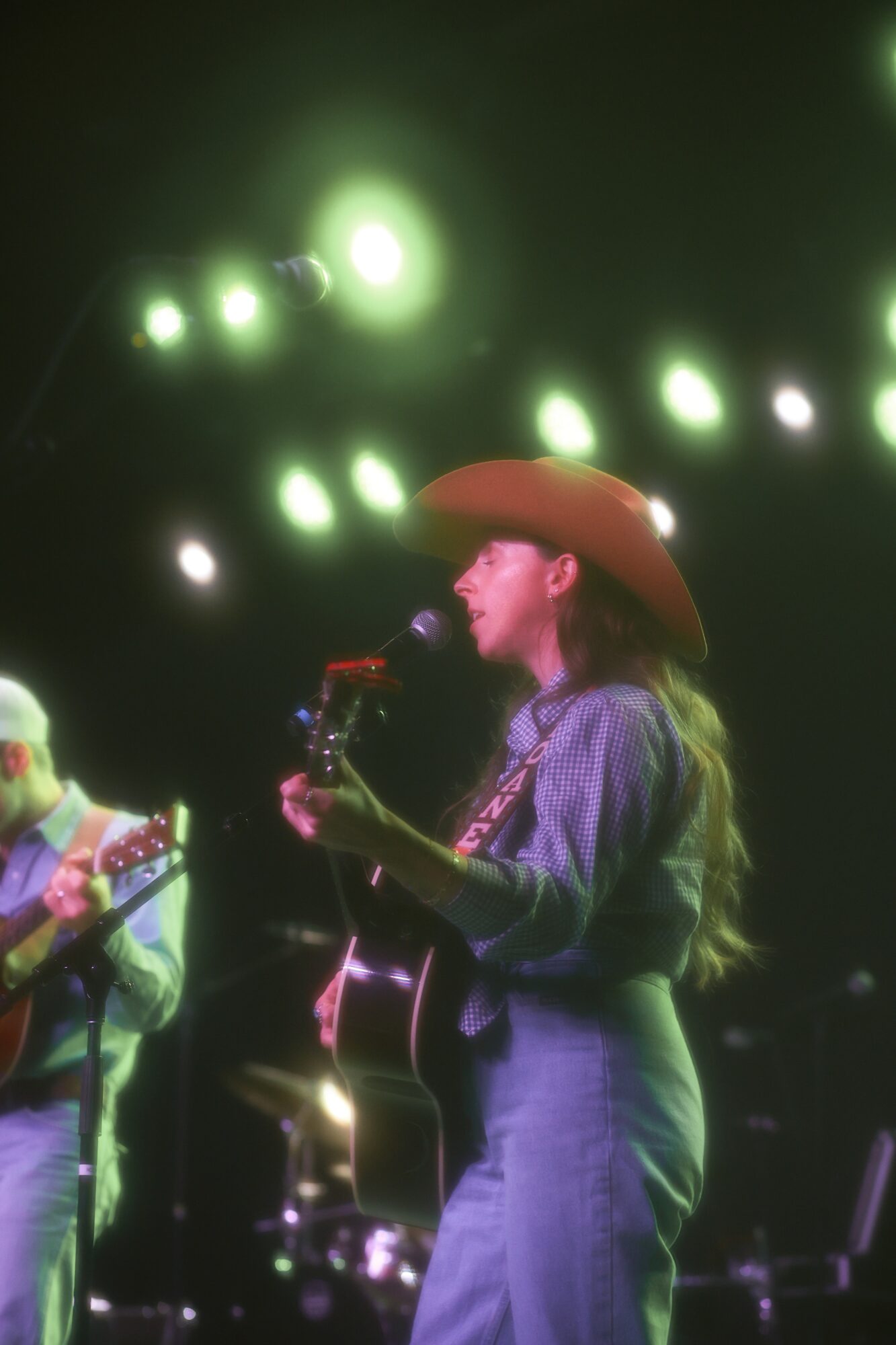 Woman wearing a cowboy hat playing guitar on stage with bright green lights overhead.