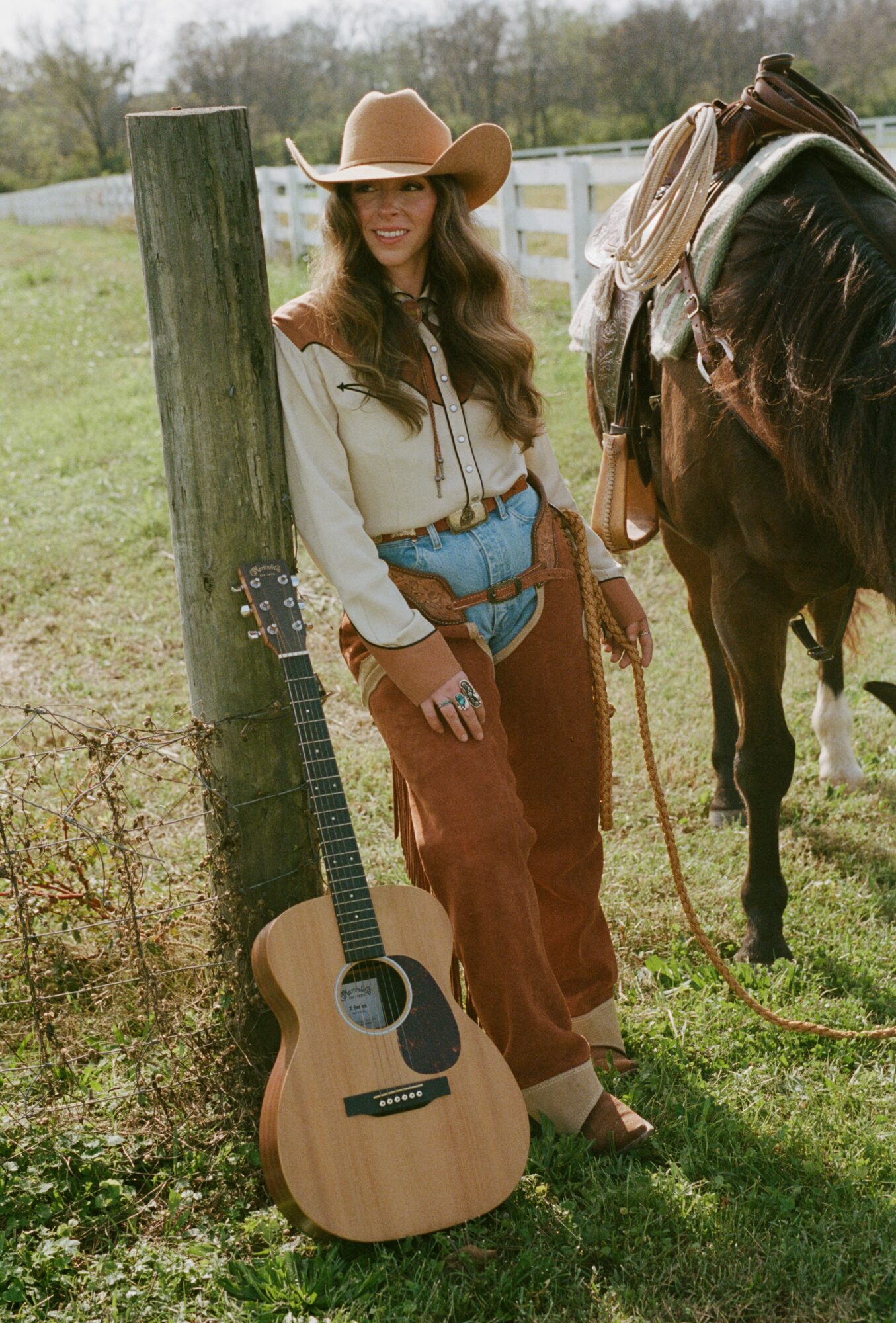 Woman in cowboy hat leaning against fence post with guitar, beside horse with saddle in outdoor setting.