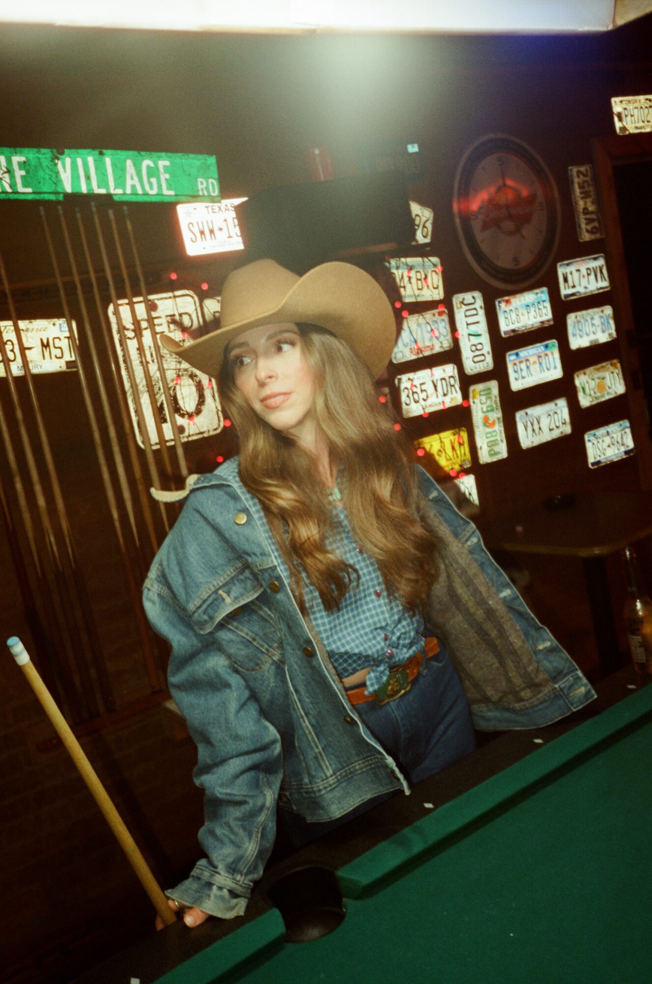 Young woman with long hair wearing a wide-brimmed hat and denim jacket, standing in a dimly lit bar with neon signs.