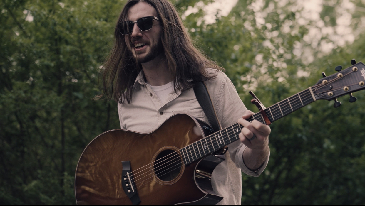 Person with long hair and sunglasses playing an acoustic guitar outdoors, smiling, surrounded by trees.