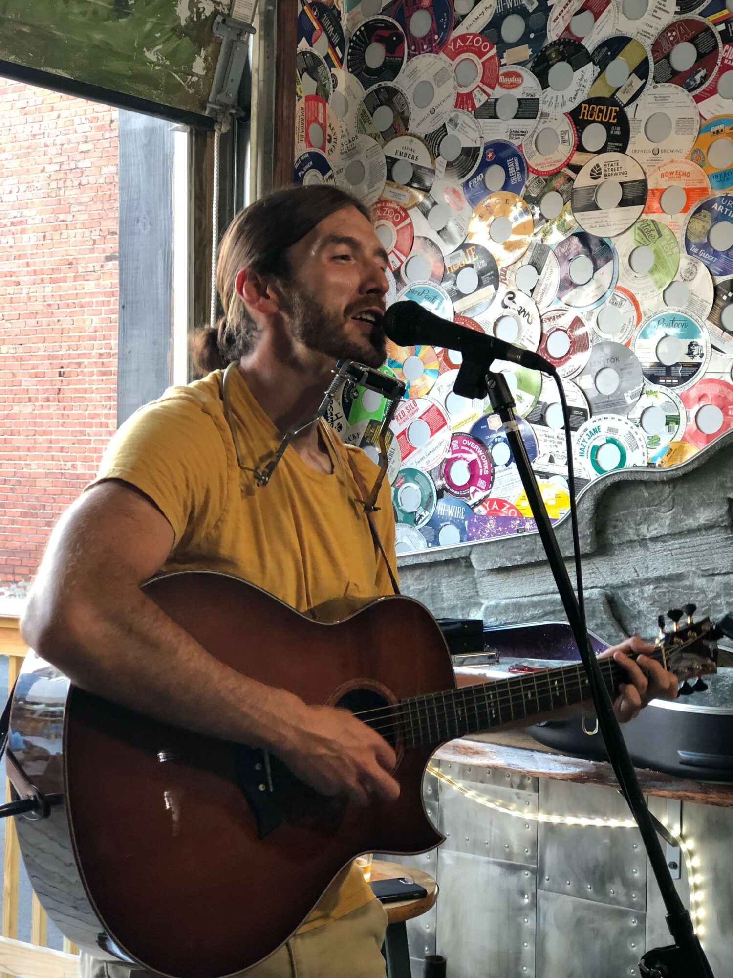 Person playing guitar and singing into microphone, colorful patterned background, indoor setting.