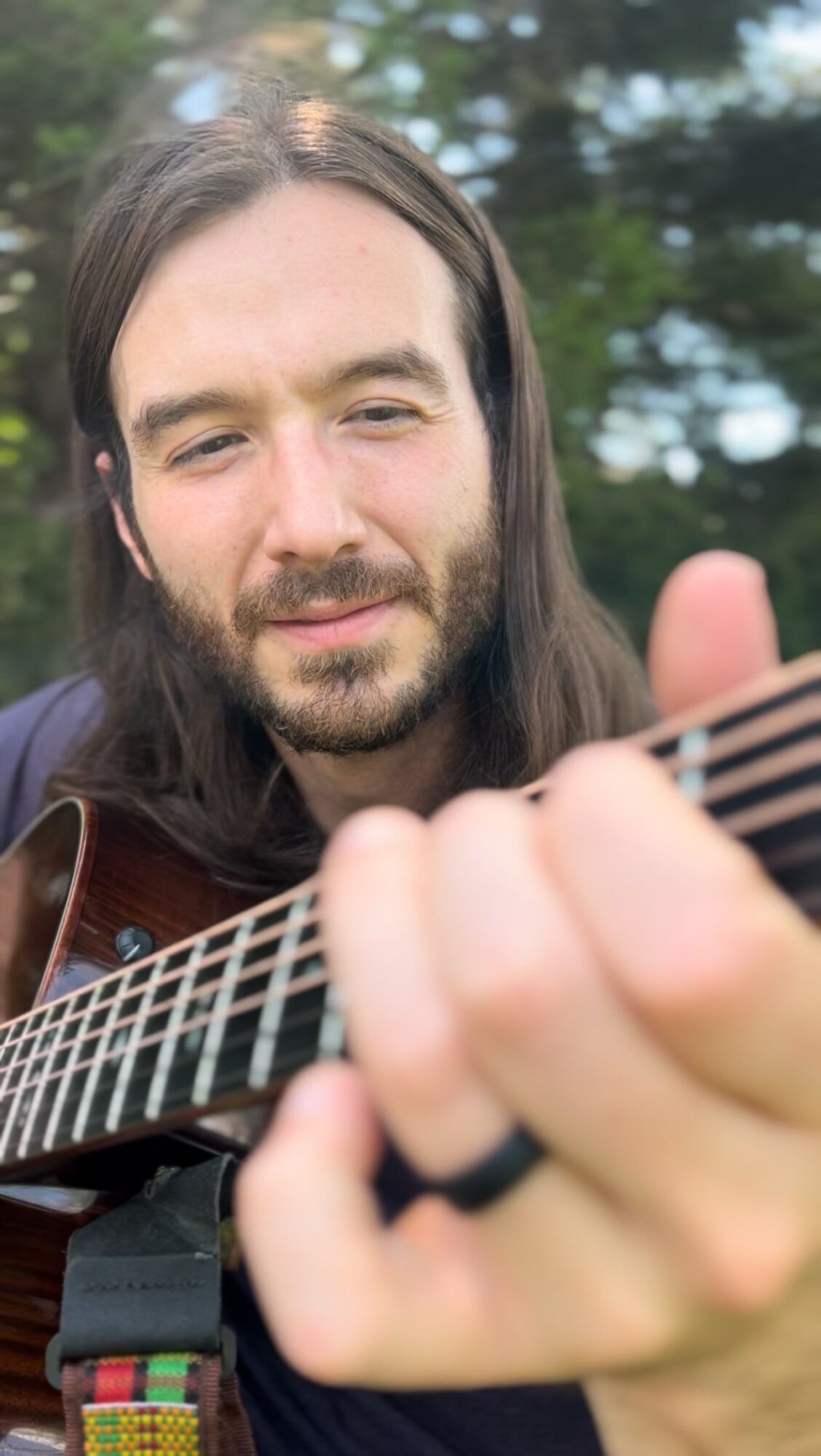Man with long hair and beard playing guitar outdoors, trees in background.