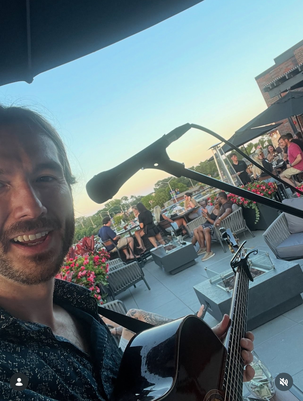 Person holding a guitar on a balcony with people and cityscape in the background.