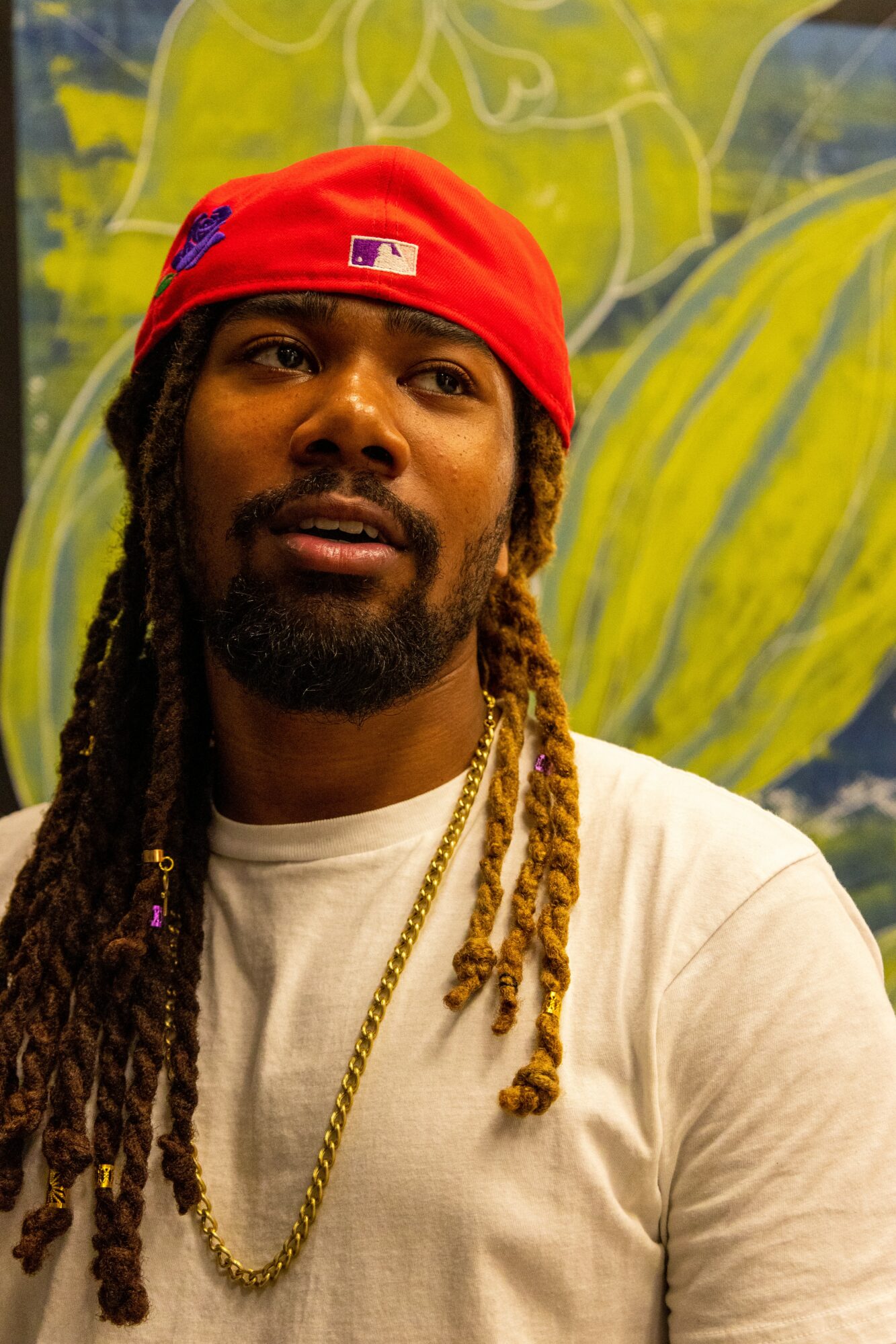 Man with long dreadlocks wearing a red cap and white shirt, standing in front of a colorful mural background.