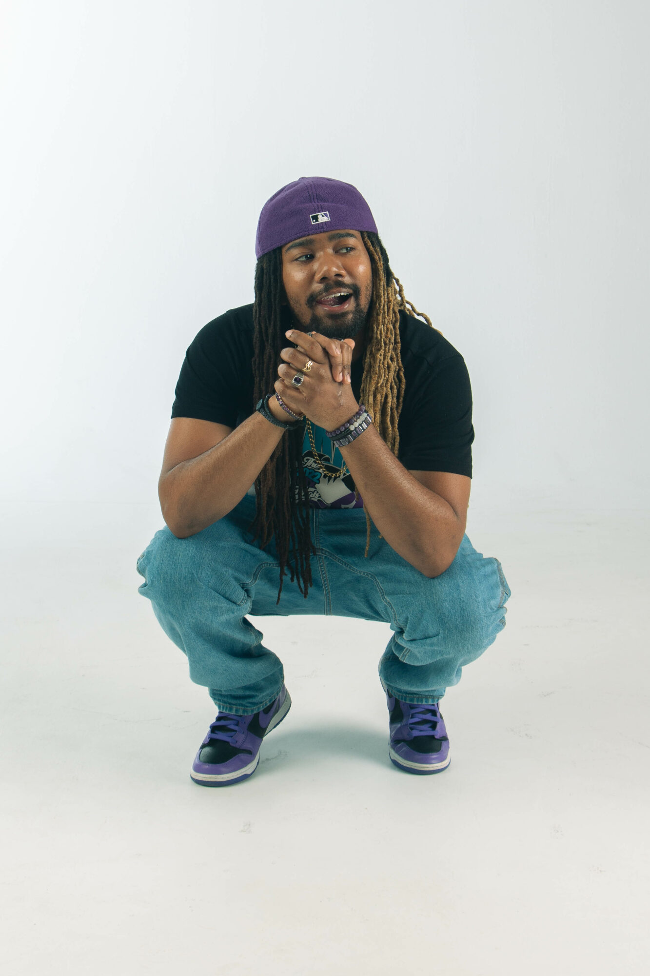 Person with long hair, purple hat, black shirt, blue jeans, squatting with hands clasped, smiling, against a plain white background.