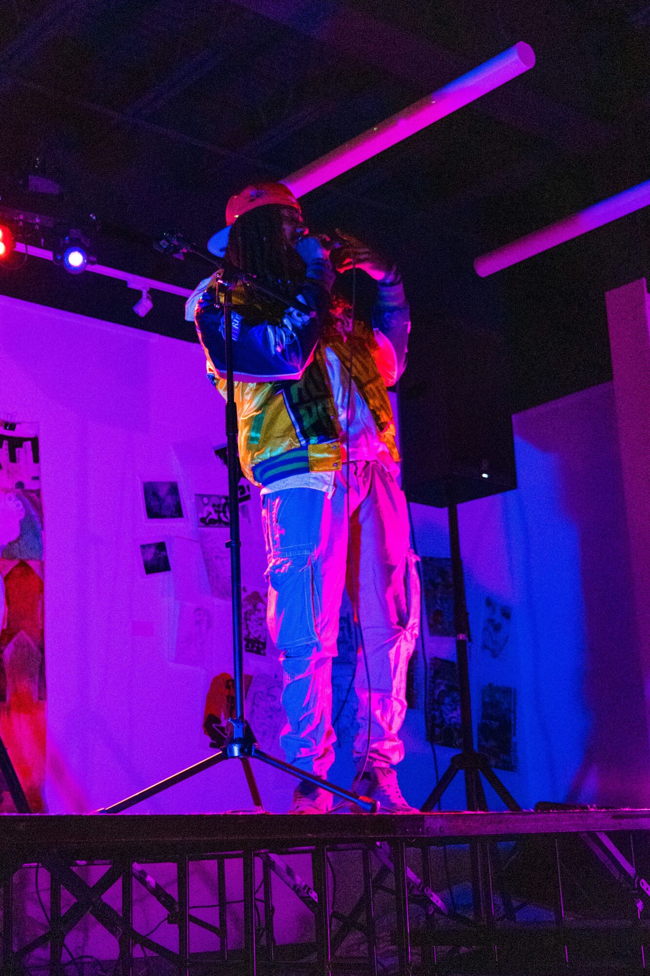 Two performers stand on stage under colorful lighting, with a microphone stand in front of them.