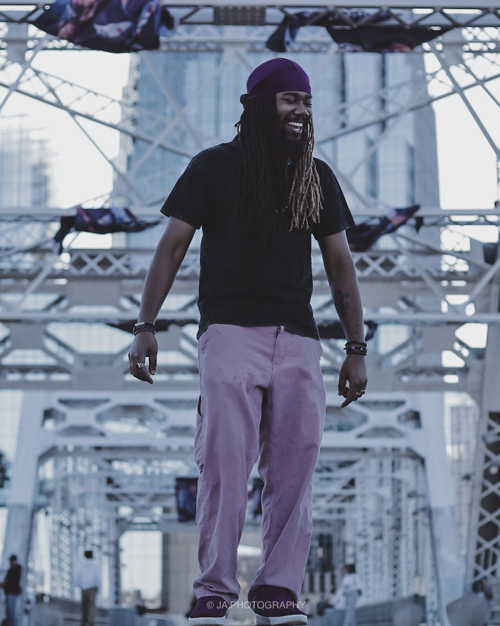 Man with long hair and beard smiling, wearing a purple beanie, black shirt, and light-colored pants, standing on a bridge.