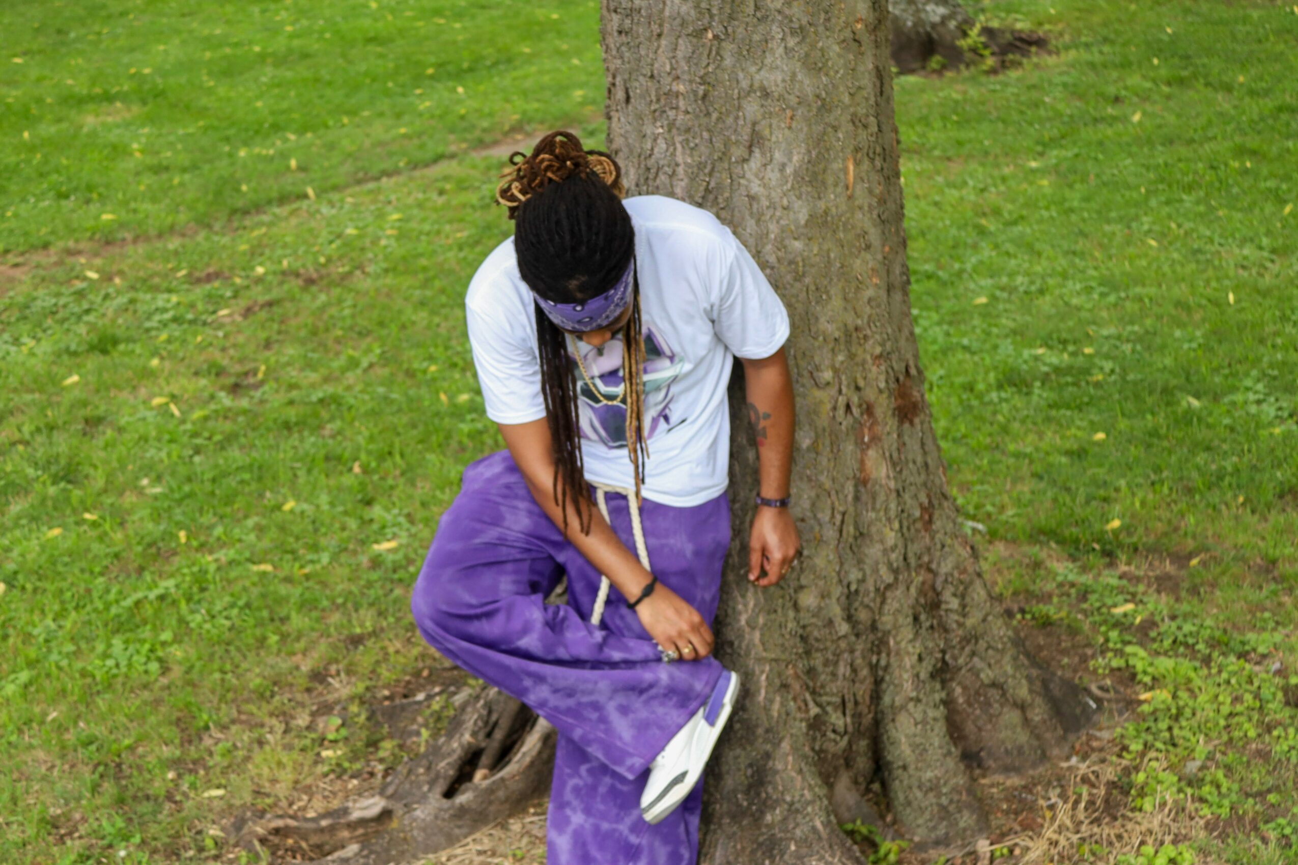 Person with long hair, wearing a white shirt and purple pants, sitting on a tree root outdoors.