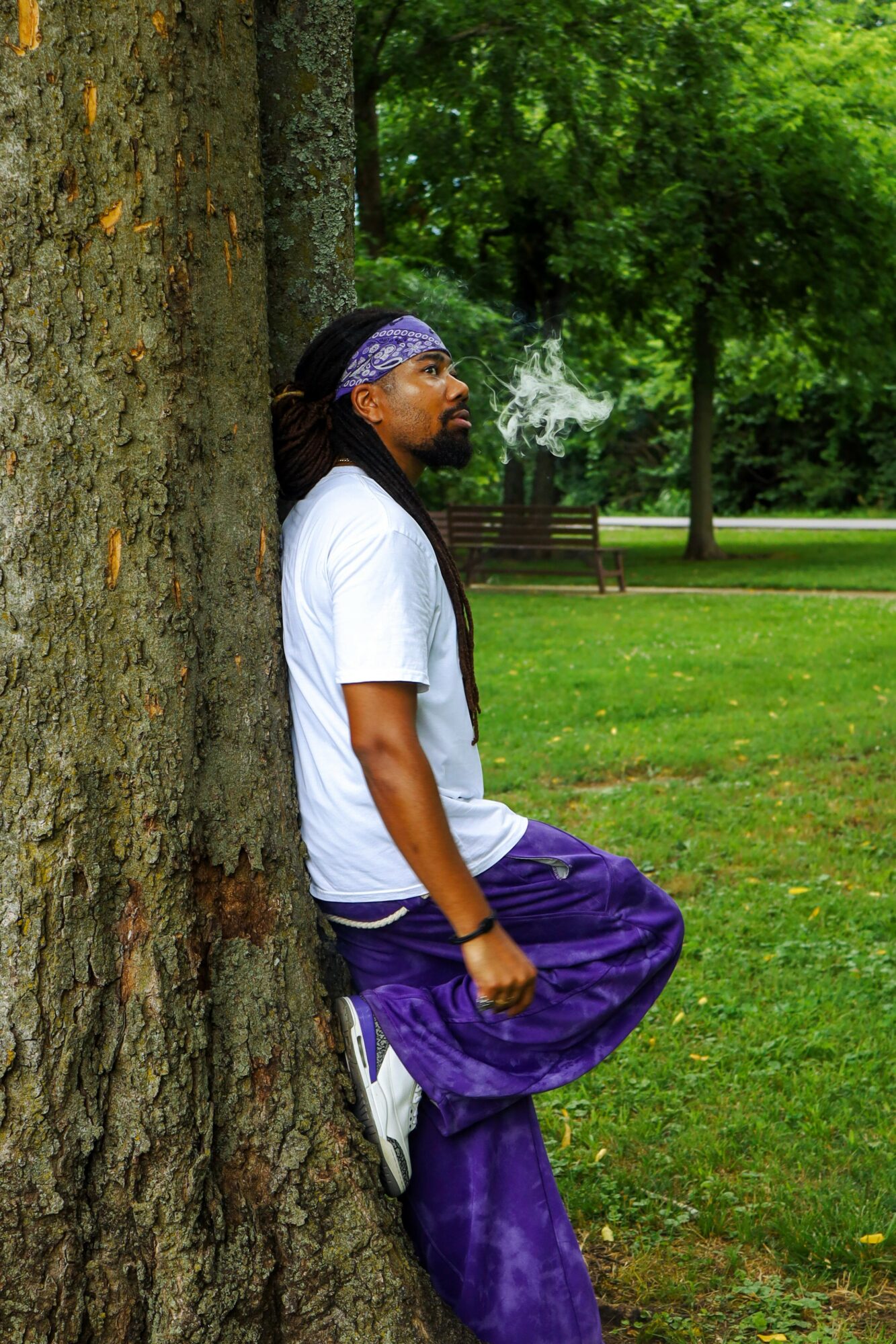 Person leaning against a tree in a park with green grass and trees, wearing a white shirt, purple pants, and a headband.