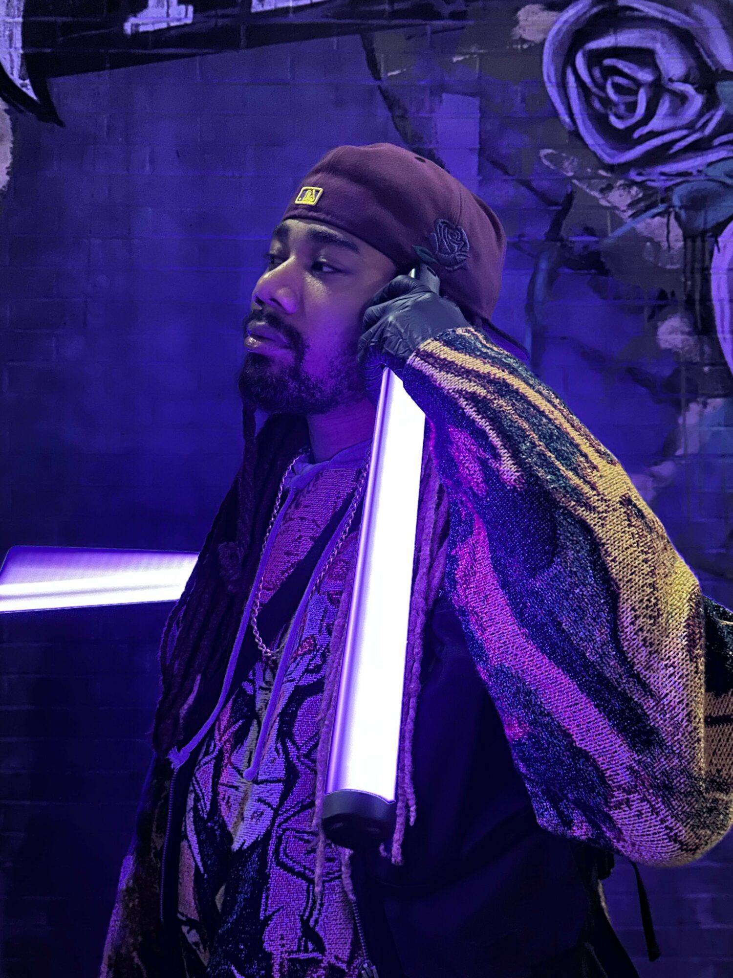 Person with short hair and patterned jacket standing in front of purple and white neon lights, with purple roses in background.