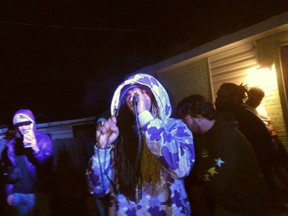 Person with long hair and a hoodie making a peace sign, surrounded by others in a dimly lit setting.